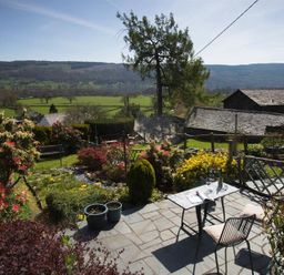 A garden with flowering bushes and a table with chairs on a stone patio overlooking fields and hills at Dormouse Cottage in Coniston