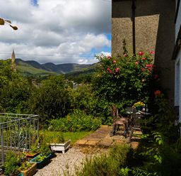 An outdoor garden area with a metal table and chairs surrounded by plants and flowers with mountains and a church steeple in the background at Uldale in Ambleside
