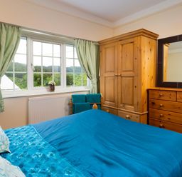 A bedroom with a bed with blue bedding a wooden wardrobe a wooden dresser with mirror and a window with green curtains at Uldale in Ambleside