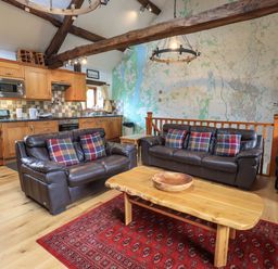A living room with leather sofas and plaid pillows a wooden coffee table a red rug and a kitchen area with wooden cabinets at Tullythwaite Garth in Crosthwaite