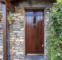A wooden door with a knocker and small window in a stone wall with a hanging flower basket lamp and greenery at Bobbin Cottage in Ings
