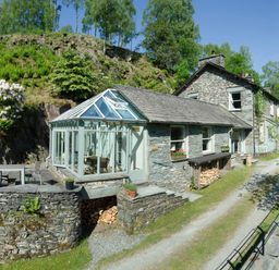 A stone house with a glass conservatory and outdoor seating area surrounded by trees and greenery at The Stable At Oakbank in Elterwater