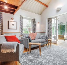 A sitting room with two gray sofas wooden side tables patterned curtains and an adjoining room with wicker chairs at The Stable At Oakbank Elterwater