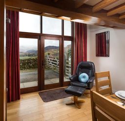 A living area with wooden dining chairs a black recliner chair with a blue cushion near glass doors with red curtains and a view of countryside hills at Nest Barn in Keswick