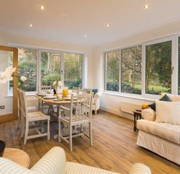A dining area with a wooden table and white chairs next to large windows and cream sofas at Lanterns At Grasmere in Grasmere