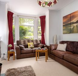A living room with brown leather sofas red curtains wooden tables and a three-panel wall art at Ellerthwaite House in Windermere