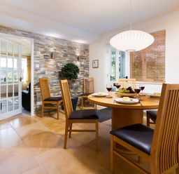 A dining room with a round wooden table set with plates glasses and a fruit bowl and wooden chairs with leather cushions at Honister Cottage in Ambleside