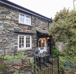 The exterior of a stone cottage with a white door and windows a small patio with a table and chairs and surrounding greenery at Cobblestone Cottage in Ambleside