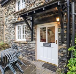 A stone cottage entrance with a white door and plastic chairs stacked on a table at Cobblestone Cottage in Ambleside