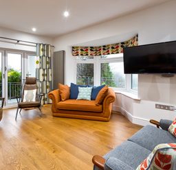 A living room with an orange sofa blue and patterned cushions a grey armchair patterned curtains and a wall-mounted TV at Wynford in Ambleside