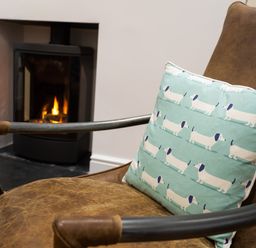 A brown leather chair with a cushion featuring white dog illustrations in front of a lit fireplace at Wynford in Ambleside