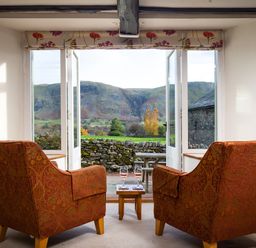 A room with two armchairs and a small table with glasses of wine facing open doors showing hills and a stone wall at Horrockwood Farm in Watermillock