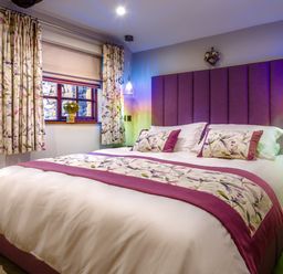 A bedroom with a purple headboard a bed with floral and purple bedding floral curtains and a window with a flower vase at Tipsy Gin Cottage in Crosthwaite