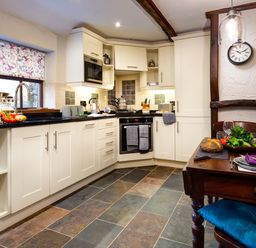 A kitchen with white cabinets a tiled floor a wooden dining table with place settings and food and a window with patterned blind at Tipsy Gin Cottage in Crosthwaite