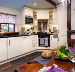 A kitchen with white cabinets a black countertop a microwave an oven and a dining table with bread and salad at Tipsy Gin Cottage in Crosthwaite