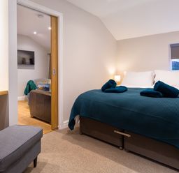 A bedroom with a bed covered with a blue blanket and rolled towels next to a window and a sliding door leading to a living area with a TV at Barn End Ambleside