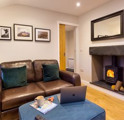 A living room with a brown leather sofa two green cushions a coffee table with a laptop a magazine and a cup a fireplace with fire and a mirror above it at Barn End Ambleside