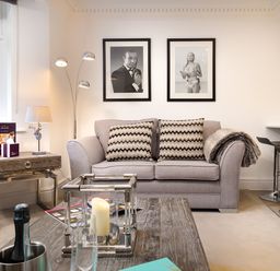 A living room with a gray sofa and two patterned cushions two black and white pictures on the wall a wooden coffee table with candles a side table with a lamp and a window in Skyfall in Bowness