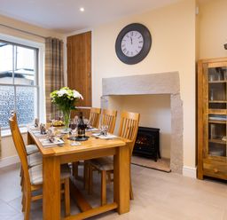 A dining room with wooden table and chairs set for dinner a clock above a fireplace and a wooden cabinet with glassware at Dovedale in Keswick