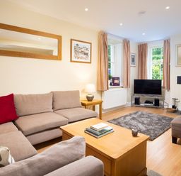A living room with a beige sectional sofa red cushions a wooden coffee table books a TV on a metal stand and windows with curtains at Scots Pine in Keswick