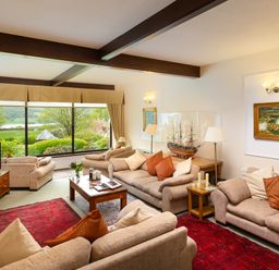 A living room with sofas, a coffee table, a model ship, framed paintings, and a large window showing a garden view at Lakefield House in Hawkshead