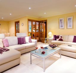 A living room with beige sofas, purple cushions, a glass coffee table with two glasses and a bowl, and a dining area with wooden furniture at Merewood Lodge in Ambleside
