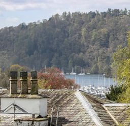 A view of rooftops with chimneys overlooking a wooded hillside and a marina with boats at The Heights in Bowness
