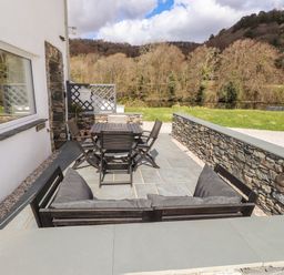 An outdoor patio with a wooden table, chairs, and a cushioned bench with a grass lawn and trees in the background at Riverside Hideaway in Newby Bridge