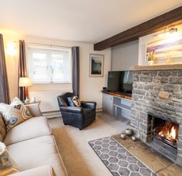 A living room with a sofa, armchair, stone fireplace with a fire, and television at Riverside Hideaway in Newby Bridge
