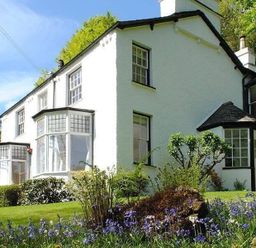 A white house with multiple windows surrounded by green bushes and purple flowers at Loughrigg Cottage in Ambleside