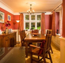 A dining room with wooden table and chairs wooden sideboard floor lamp and paintings on orange walls at Priory Lodge in Windermere