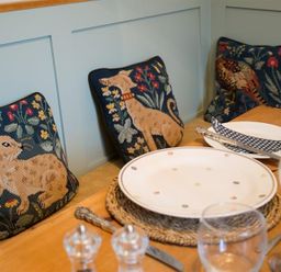 A dining table set with plates glasses and cutlery next to cushions with animal designs on a bench at The Blithe Hare in Troutbeck Bridge