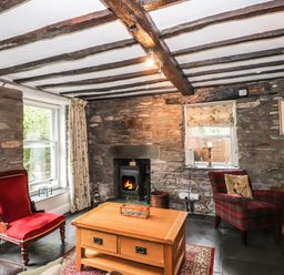 A living room with stone walls a wood burning stove wooden beams on the ceiling and wooden furniture at The Mews in Ambleside