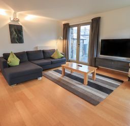 A living room with a dark sofa green cushions wooden coffee table a striped rug a wall-mounted television and large windows with curtains at Idle Mill 1 Ambleside