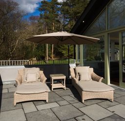 Two patio loungers with cushions under a large umbrella on a stone terrace outside a building with glass doors at Tall Trees in Bowness