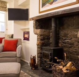 A sitting room with a stone fireplace and wood stove a basket of firewood an armchair with a red cushion and a wall-mounted television at Lettera in Ambleside