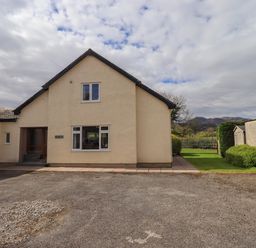 A cream house with a gravel driveway and a small garden with trees and mountains in the background at Laal Field House in Keswick