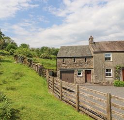 A stone and stucco house with wooden doors next to a grassy hill and wooden fence at Goswick Hall in Bowland Bridge