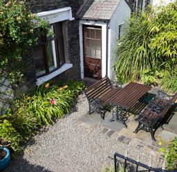 A garden patio with wooden table and benches surrounded by plants and a stone house at Honeysuckle Cottage in Bowness