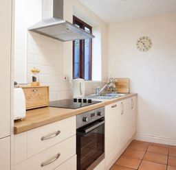 A kitchen with wooden countertop white cabinets an oven a stove a kettle a bread box and a wall clock at Honeysuckle Cottage in Bowness