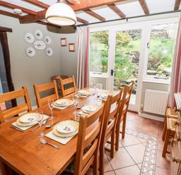 A dining room with a wooden table set for six near windows with curtains at Gunpowder Cottage in Langdale