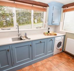 A kitchen with blue cabinets a stainless steel sink a washing machine and a window showing stone wall at Gunpowder Cottage in Langdale