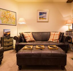 A living room with leather sofa and armchair with plaid pillows a large ottoman with trays and remote controls lamps framed pictures and a bookshelf at Orrest Range in Windermere