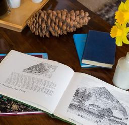 A wooden table with an open book showing sketches and text pine cone two closed books and a vase with yellow flowers at Camellia Cottage in Bowness