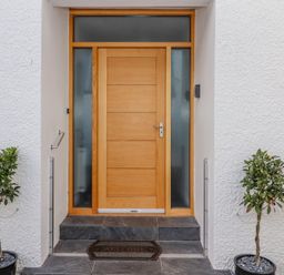 A wooden front door with frosted glass panels on a white textured wall with two potted plants on either side and a welcome mat at Bowderstone in Keswick