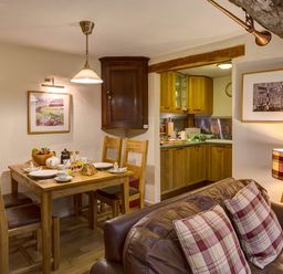 A dining area with a wooden table and chairs set for breakfast next to a kitchen with wooden cabinets and a living area with a sofa and plaid cushions at Burrow Cottage in Bowness