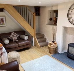 A living room with a leather sofa under stairs a fireplace with firewood and a large clock on the wall at Burrow Cottage in Bowness