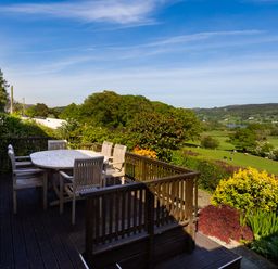 An outdoor wooden deck with a table and chairs overlooking a green field and distant hills at Garden Cottage At Coniston in Coniston