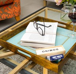 A glass coffee table with a wooden tray holding an open book with glasses a domino set a deck of playing cards and a glass of red wine near an orange patterned chair
