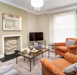 A living room with orange patterned armchairs a glass coffee table a television a mirror above a fireplace and white blinds at Pippin Cottage in Keswick